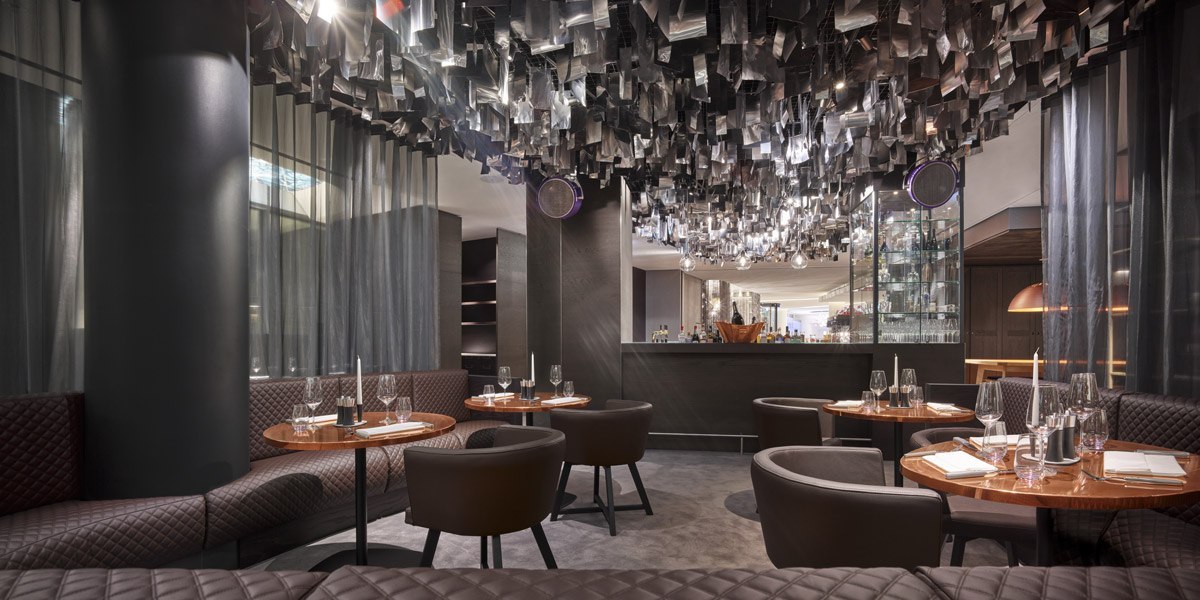 Elegantes Restaurant mit runden Tischen, gepolsterten Stühlen und einer modernen Bar. Die Decke ist mit dekorativen Elementen verziert., © Jaz Hotel Stuttgart