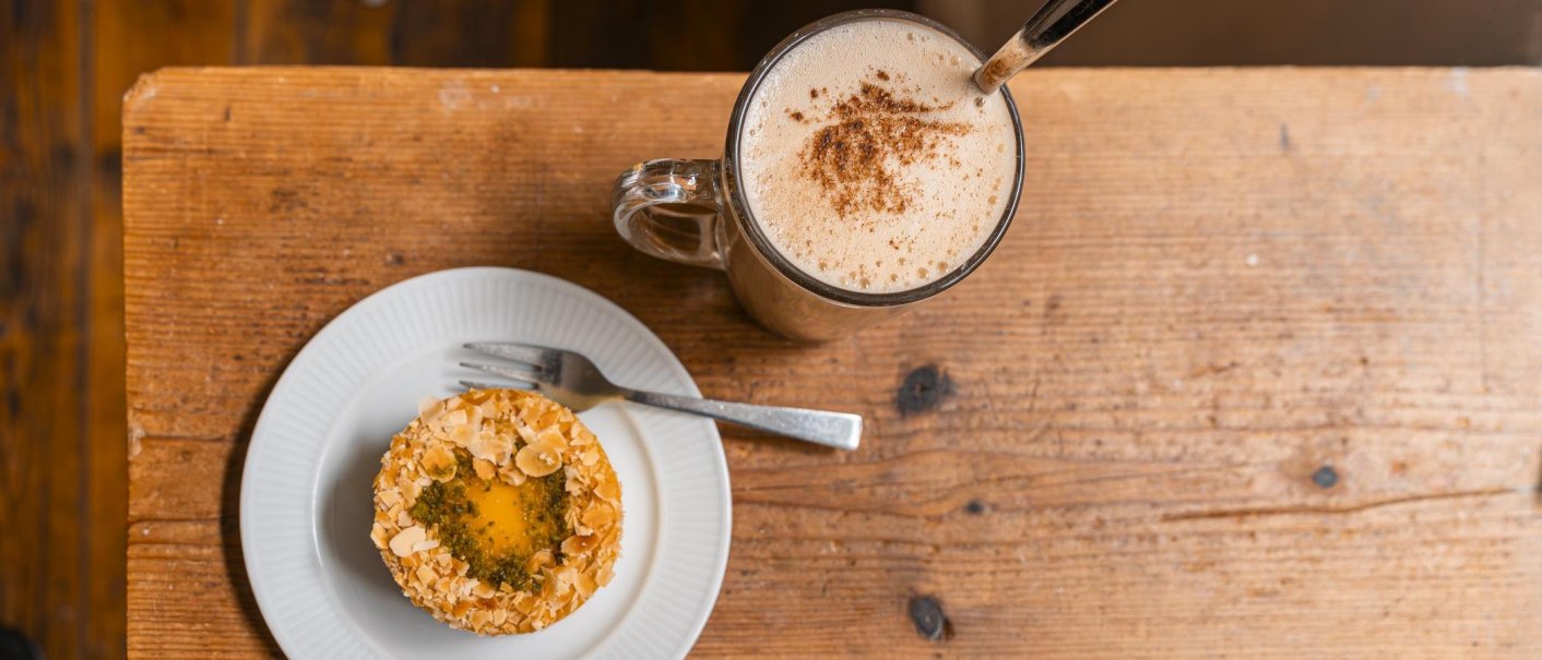 Ein Teller mit einem Gebäck, garniert mit Mandeln, und eine Tasse Kaffee mit Milchschaum auf einem rustikalen Holztisch., © SMG, Sarah Schmid