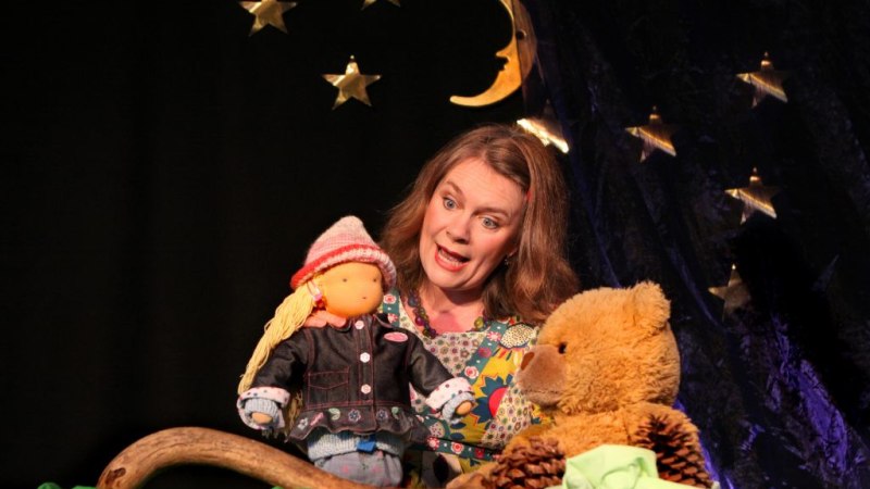 Eine Frau interagiert mit einer Puppe und einem Teddybär vor einem sternen- und mondgeschmückten Hintergrund., © Theater Hammerschmiede Eine Frau interagiert mit einer Puppe und einem Teddybär vor einem sternen- und mondgeschmückten Hintergrund., © Theater Hammerschmiede