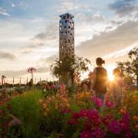 Ein Turm im Himmelsgarten, umgeben von bunten Blumen. Zwei Personen stehen im Vordergrund, während die Sonne im Hintergrund untergeht., © Foto Thomas Zehnder