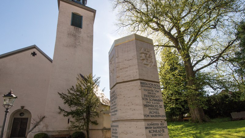 Die Evangelische Stiftskirche Bad Boll mit einem Denkmal im Vordergrund, umgeben von Bäumen und einer Laterne., © SMG, Achim Mende