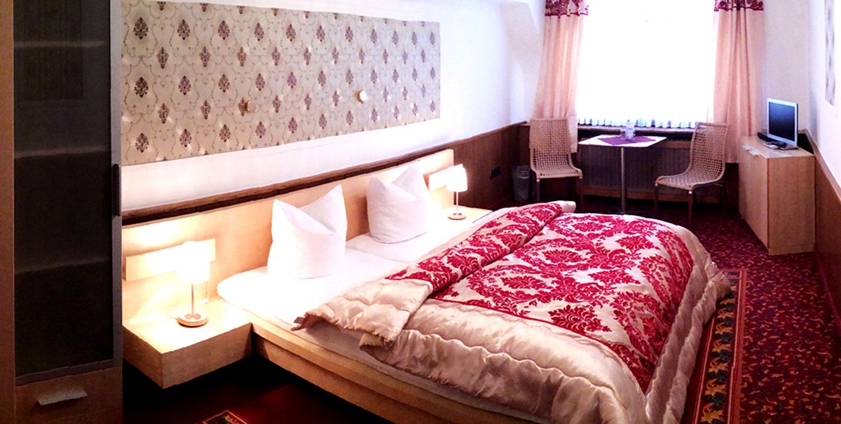 Gemütliches Hotelzimmer mit Doppelbett, rotem Teppich, Vorhängen, kleinem Tisch, zwei Stühlen und Fernseher. Gemütliches Hotelzimmer mit Doppelbett, rotem Teppich, Vorhängen, kleinem Tisch, zwei Stühlen und Fernseher.