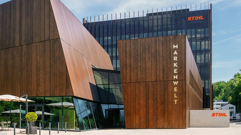 Moderne Gebäude der STIHL Markenwelt mit Holzverkleidung und Glasfronten, umgeben von Bäumen. Sonnenschirme und Sitzgelegenheiten im Vordergrund., © STIHL Markenwelt