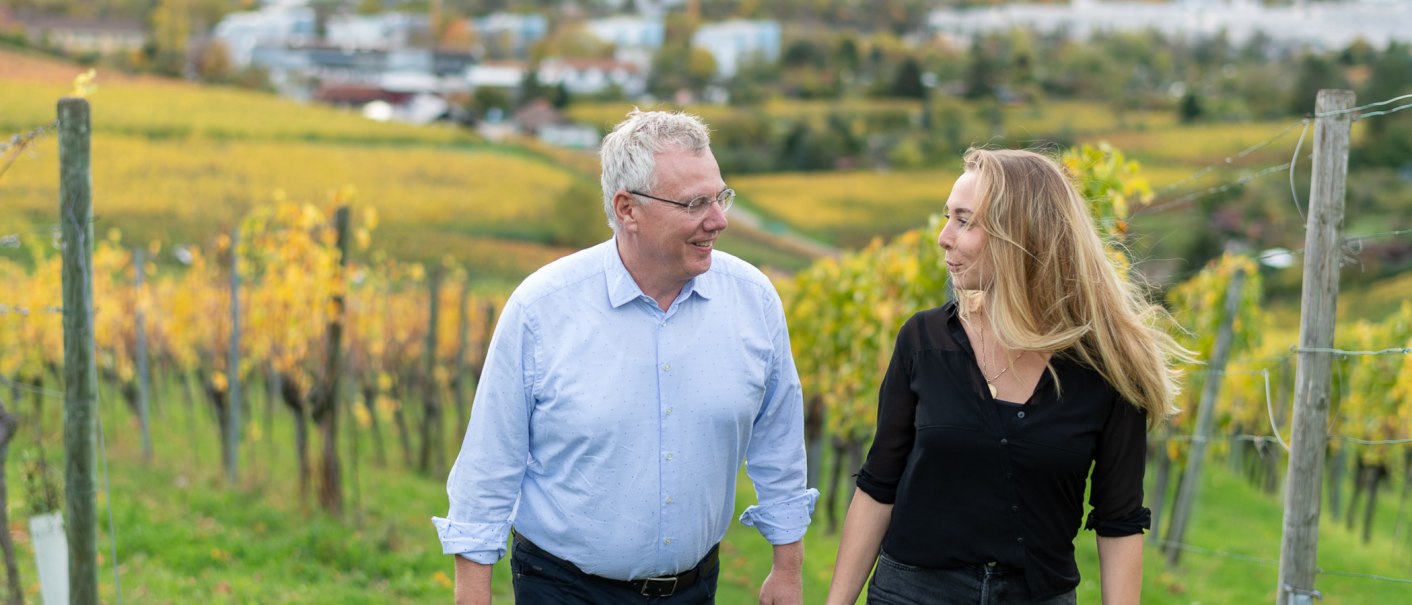 Zwei Personen spazieren lächelnd durch einen herbstlichen Weinberg. Im Hintergrund sind gelbe Weinreben und eine unscharfe Landschaft zu sehen., © BURKHARDT HELLWIG
