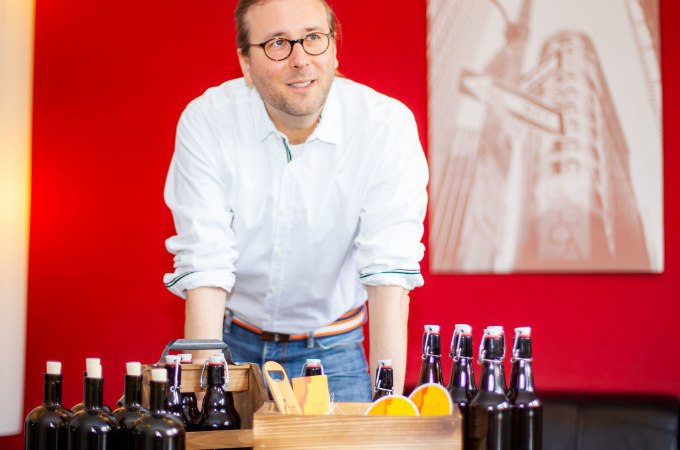 Ein Mann mit Brille pr&auml;sentiert verschiedene Bierflaschen und eine Holzkiste mit Orangen vor einem roten Hintergrund., &copy; PlochingenInfo