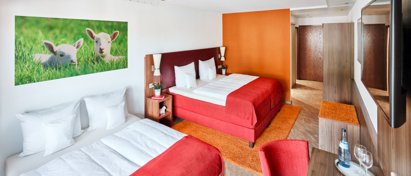Modernes Hotelzimmer mit zwei großen Betten, roter Bettwäsche und oranger Wand. Ein Bild von Lämmern hängt an der Wand. Ein Tisch mit Wasserflasche und Gläsern., © Hotel Restaurant Lamm
