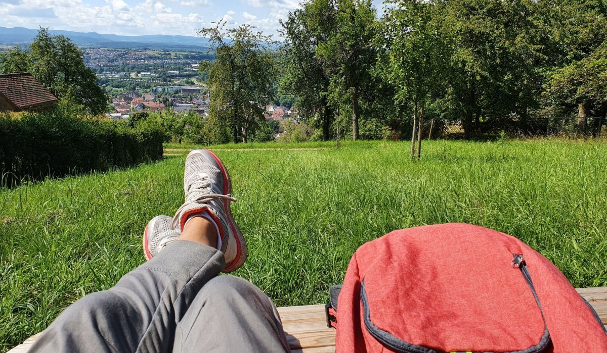 Person entspannt auf einer Bank mit Blick auf eine Stadt in der Ferne, umgeben von grünen Wiesen und Bäumen. Ein roter Rucksack liegt daneben. Person entspannt auf einer Bank mit Blick auf eine Stadt in der Ferne, umgeben von grünen Wiesen und Bäumen. Ein roter Rucksack liegt daneben.