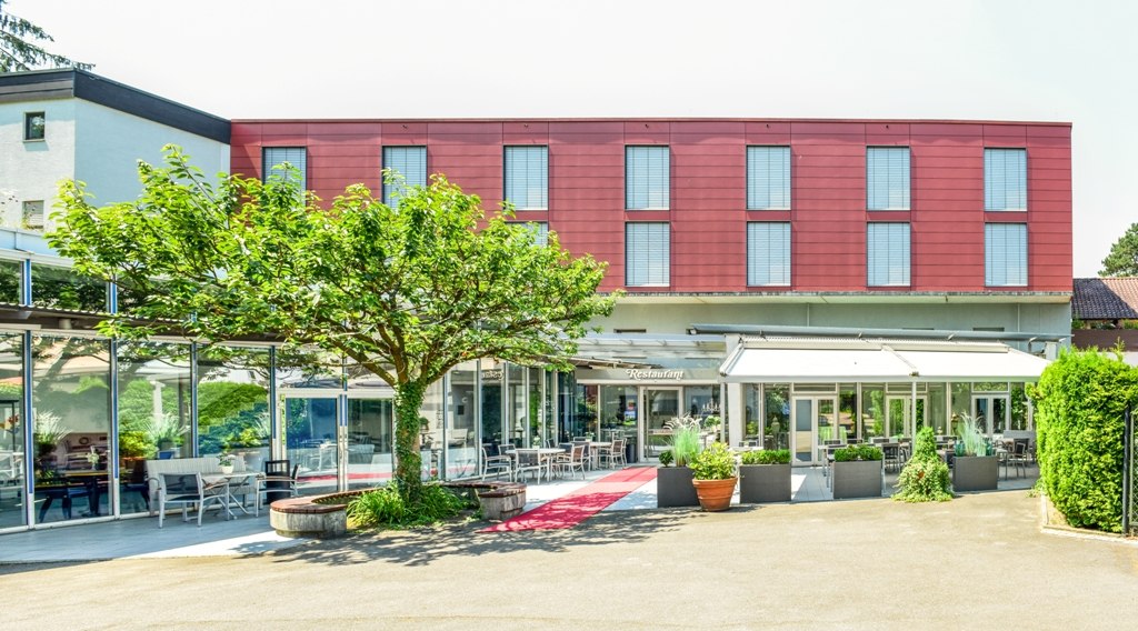 Modernes Gebäude mit roter Fassade und Glasfront, umgeben von Pflanzen und Terrasse mit Sitzgelegenheiten., © Hotel Fuchsen Modernes Gebäude mit roter Fassade und Glasfront, umgeben von Pflanzen und Terrasse mit Sitzgelegenheiten., © Hotel Fuchsen