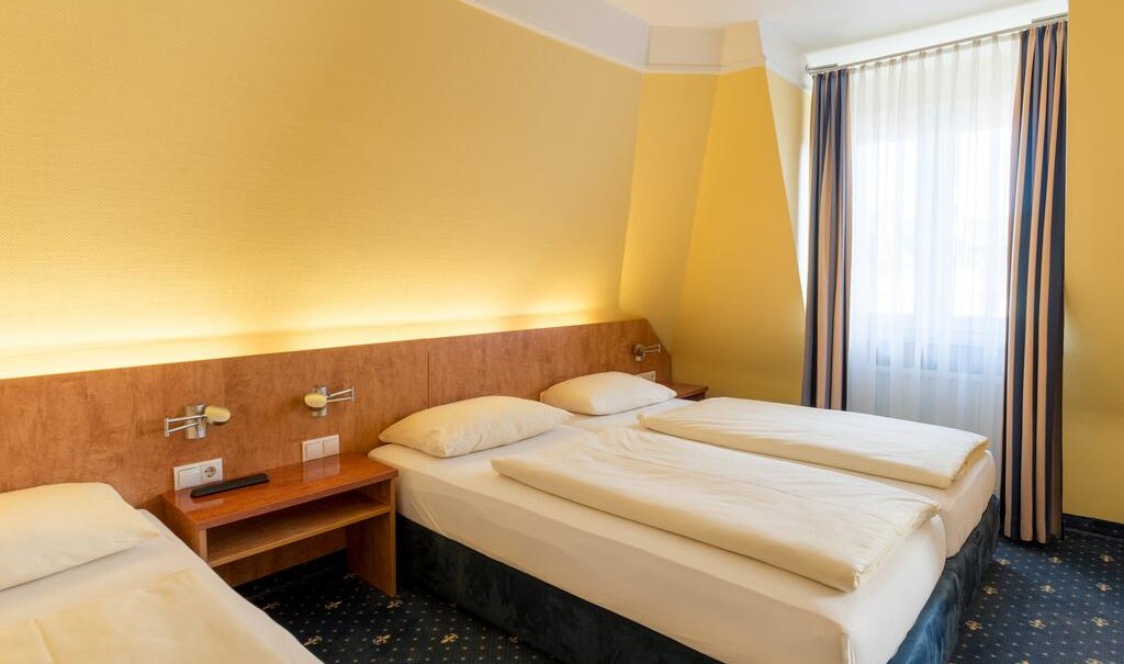 Ein Hotelzimmer mit zwei Einzelbetten, gelben Wänden und blau-gemustertem Teppich. Ein Fenster mit gestreiften Vorhängen lässt Licht herein., © Aparthotel Astro