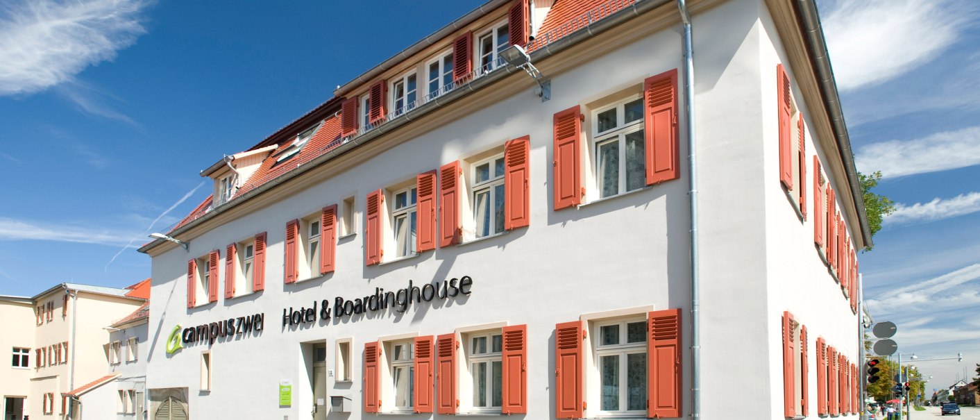 Weißes Gebäude mit roten Fensterläden, blauer Himmel. Schriftzug: 'campuszwei Hotel & Boardinghouse'. Moderne Architektur, sonniger Tag., © TOMAS