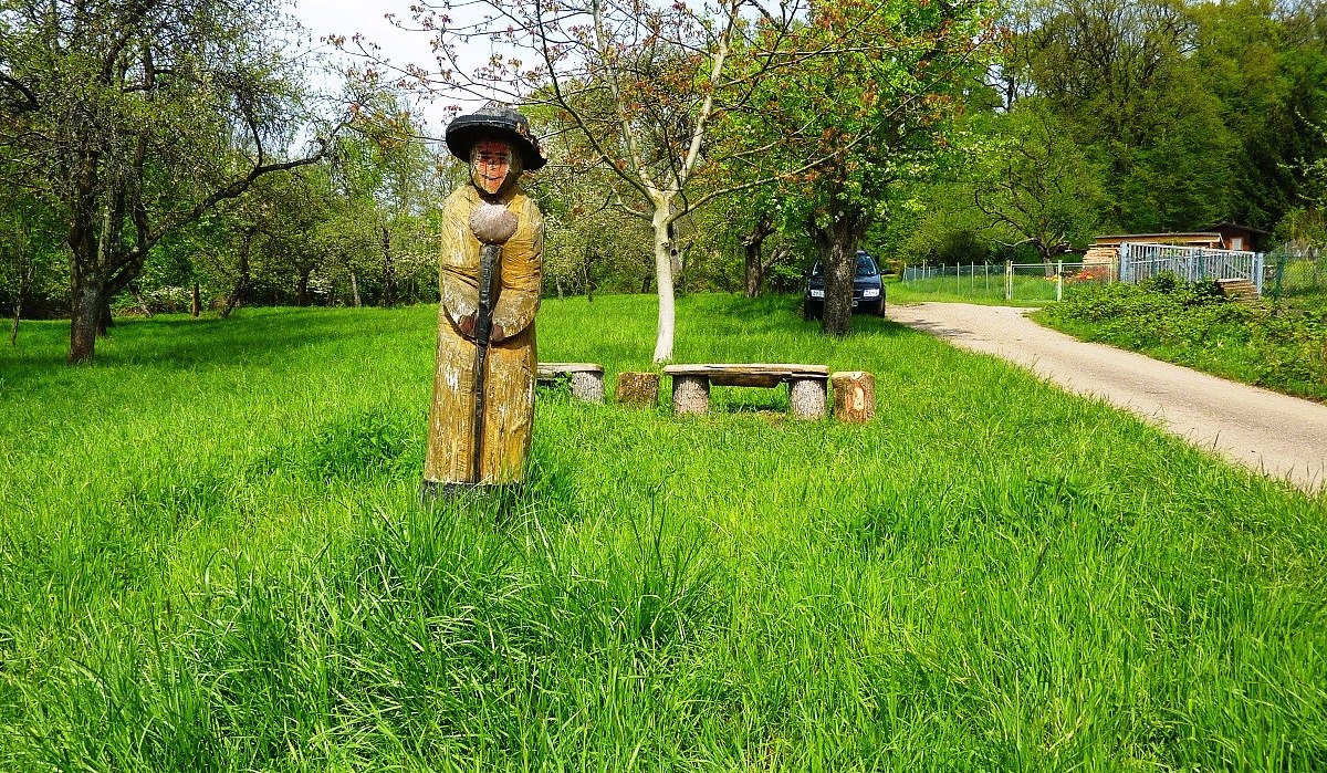 Holzfigur eines Pilgers auf einer grünen Wiese, umgeben von Bäumen. Ein Weg und eine Bank sind im Hintergrund zu sehen., © Plochingen