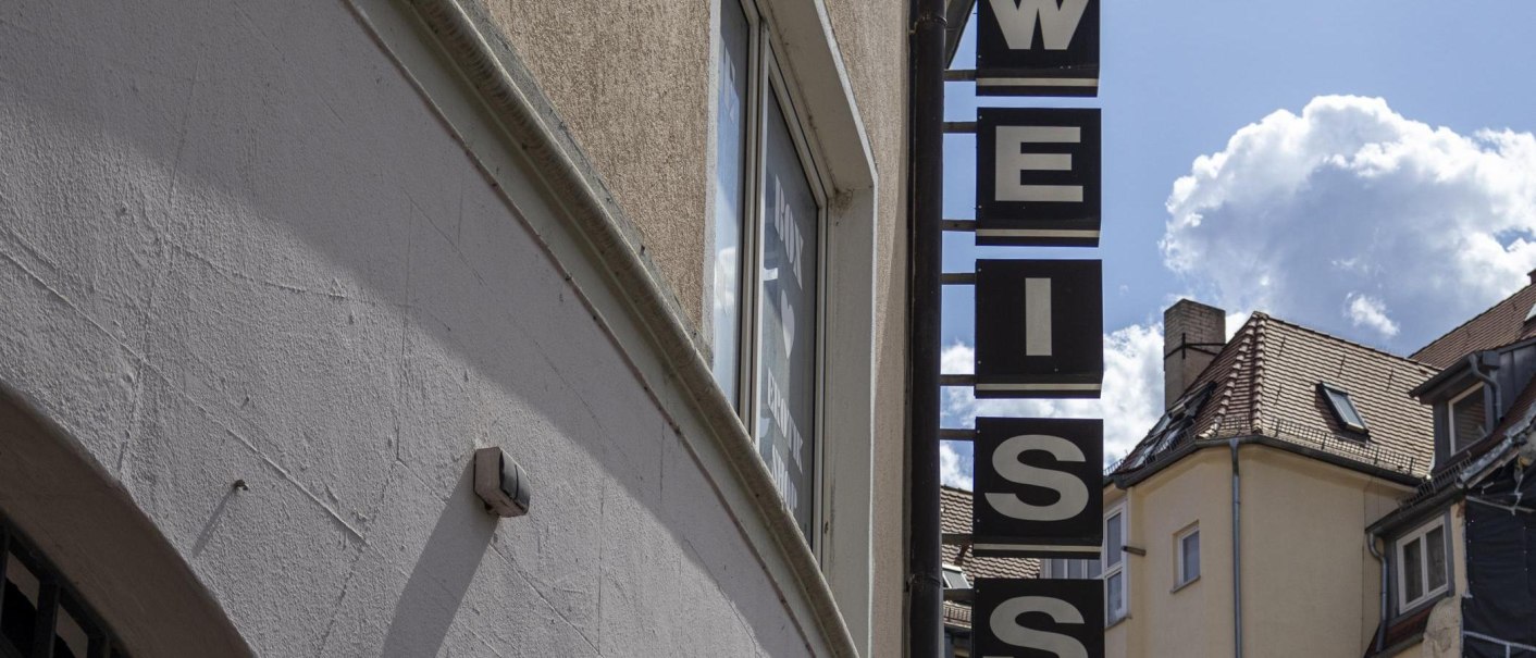 Ein Schild mit der Aufschrift 'WEISS' hängt an einem Gebäude. Der Himmel ist blau mit einigen Wolken. Im Hintergrund sind Dächer zu sehen., © SMG, Sarah Schmid Ein Schild mit der Aufschrift 'WEISS' hängt an einem Gebäude. Der Himmel ist blau mit einigen Wolken. Im Hintergrund sind Dächer zu sehen., © SMG, Sarah Schmid