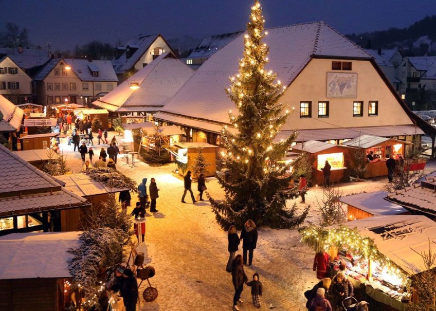 Verschneiter Weihnachtsmarkt in Metzingen mit beleuchtetem Baum und Ständen. Menschen schlendern durch die festliche Atmosphäre auf dem Kelternplatz., © Stadt Metzingen
