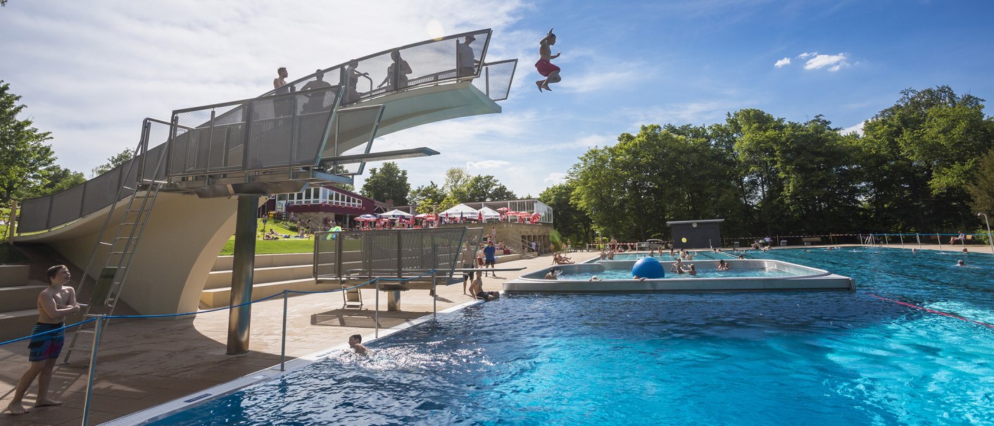 Ein Freibad mit Sprungturm, von dem ein Mensch ins Wasser springt. Viele Menschen schwimmen im großen Becken. Im Hintergrund sind Bäume und Sonnenschirme., © Stuttgarter Bäder Ein Freibad mit Sprungturm, von dem ein Mensch ins Wasser springt. Viele Menschen schwimmen im großen Becken. Im Hintergrund sind Bäume und Sonnenschirme., © Stuttgarter Bäder