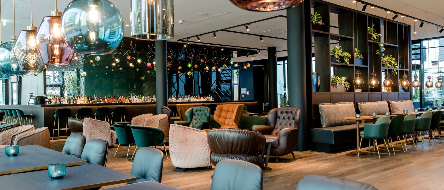 Ein modernes Restaurant mit eleganten Hängelampen, gemütlichen Sesseln und einer Bar im Hintergrund. Pflanzen dekorieren die Regale., © Motel One - Stuttgart Hauptbahnhof