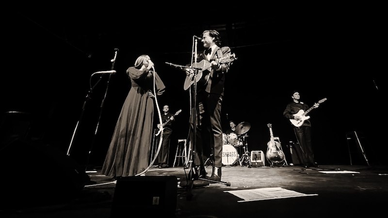 Schwarz-weiß Bild einer Band auf der Bühne. Sängerin und Sänger mit Gitarre im Vordergrund, Schlagzeuger und Gitarristen im Hintergrund., © Good Concert GmbH Schwarz-weiß Bild einer Band auf der Bühne. Sängerin und Sänger mit Gitarre im Vordergrund, Schlagzeuger und Gitarristen im Hintergrund., © Good Concert GmbH