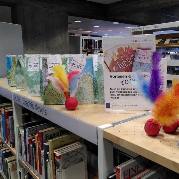 Regal in Bibliothek mit Bastelsets und Schild 'Vorlesen & Basteln to go'. Bunte Federn und Bücher im Hintergrund., © Stadtbibliothek Sindelfingen