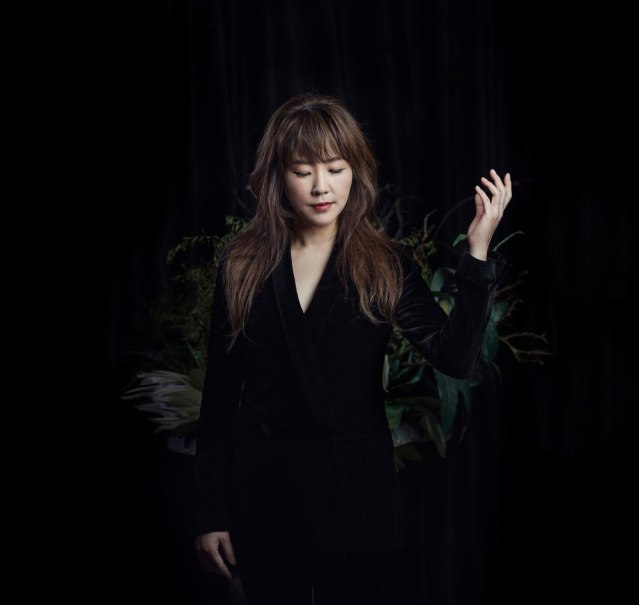 Eine Frau in schwarzer Kleidung steht vor einem dunklen Hintergrund mit Pflanzen. Sie hat die Augen geschlossen und hebt eine Hand leicht an., &copy; Youn Sun Nah &copy; Seung Yull Nah