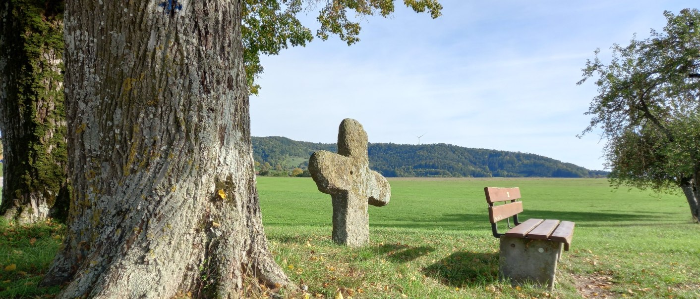Ein S&uuml;hnekreuz neben einer Bank unter einem Baum auf einer gr&uuml;nen Wiese. Im Hintergrund sind H&uuml;gel und ein Windrad zu sehen., &copy; Petra Natzkowski