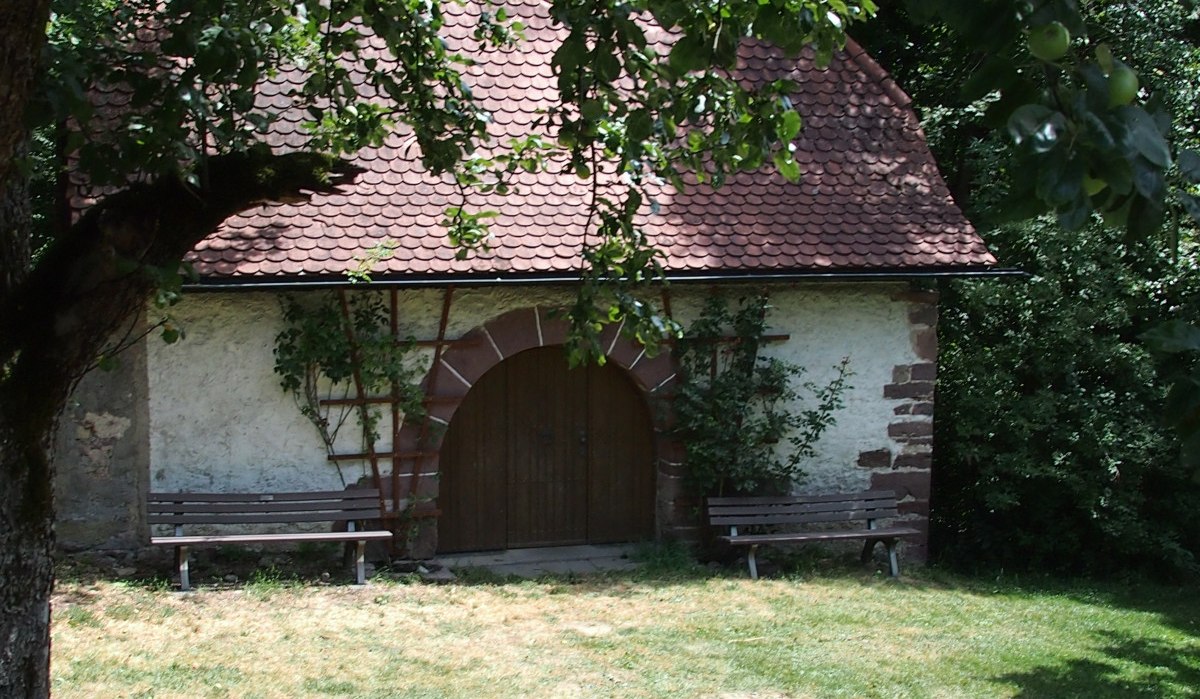 Kleine Hütte mit rotem Ziegeldach und Holztür, umgeben von Bäumen und Bänken im Grünen., © Natur.Nah. Schönbuch & Heckengäu Kleine Hütte mit rotem Ziegeldach und Holztür, umgeben von Bäumen und Bänken im Grünen., © Natur.Nah. Schönbuch & Heckengäu