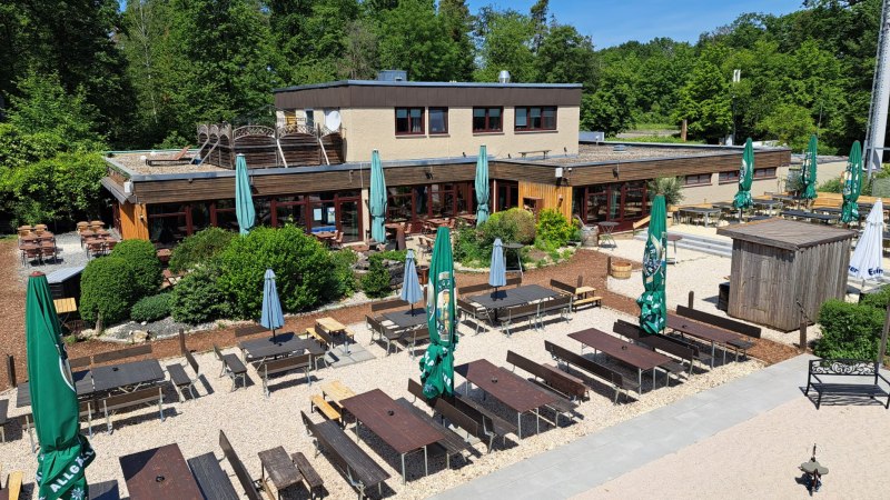 Außenbereich von Weitmanns Waldhaus mit Holztischen, Bänken und Sonnenschirmen, umgeben von grünen Bäumen und blauen Himmel., © Timo Weitmann Außenbereich von Weitmanns Waldhaus mit Holztischen, Bänken und Sonnenschirmen, umgeben von grünen Bäumen und blauen Himmel., © Timo Weitmann