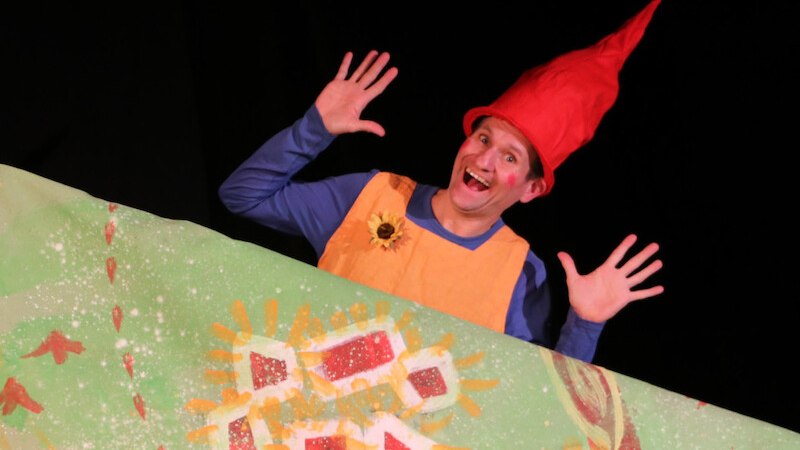 Ein Schauspieler in einem Zwergenkostüm mit roter Mütze und orangefarbenem Oberteil lächelt und hebt die Hände vor einem bunten Hintergrund., © Kultur- und Sportamt Stadt Bietigheim-Bissingen