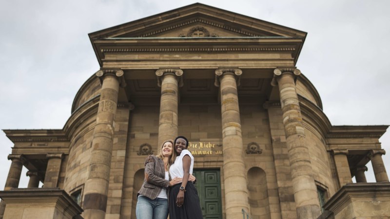 Zwei Personen umarmen sich lächelnd vor der imposanten Fassade der Grabkapelle Stuttgart mit ihren Säulen., © Stuttgart-Marketing GmbH, wpsteinheisser