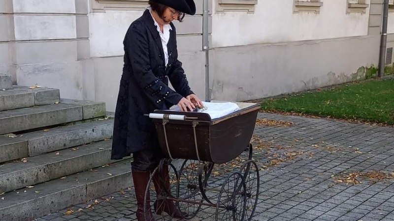 Eine Person in historischer Kleidung steht neben einem antiken Kinderwagen auf einem gepflasterten Weg vor einem Geb&auml;ude mit wei&szlig;en Fenstern., &copy; Margit Sch&auml;fer