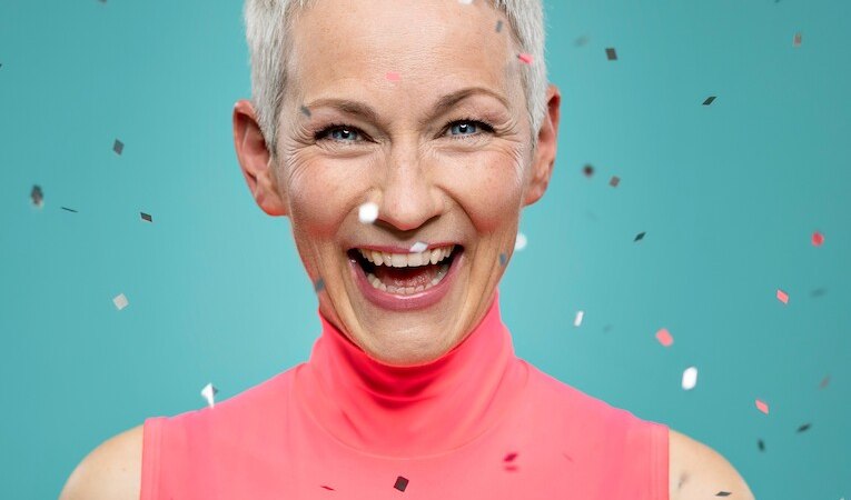 Eine lachende Frau mit kurzen grauen Haaren und einem rosa Oberteil ist von Konfetti umgeben. Der Hintergrund ist blau., &copy; Kulturreferat Bad Urach