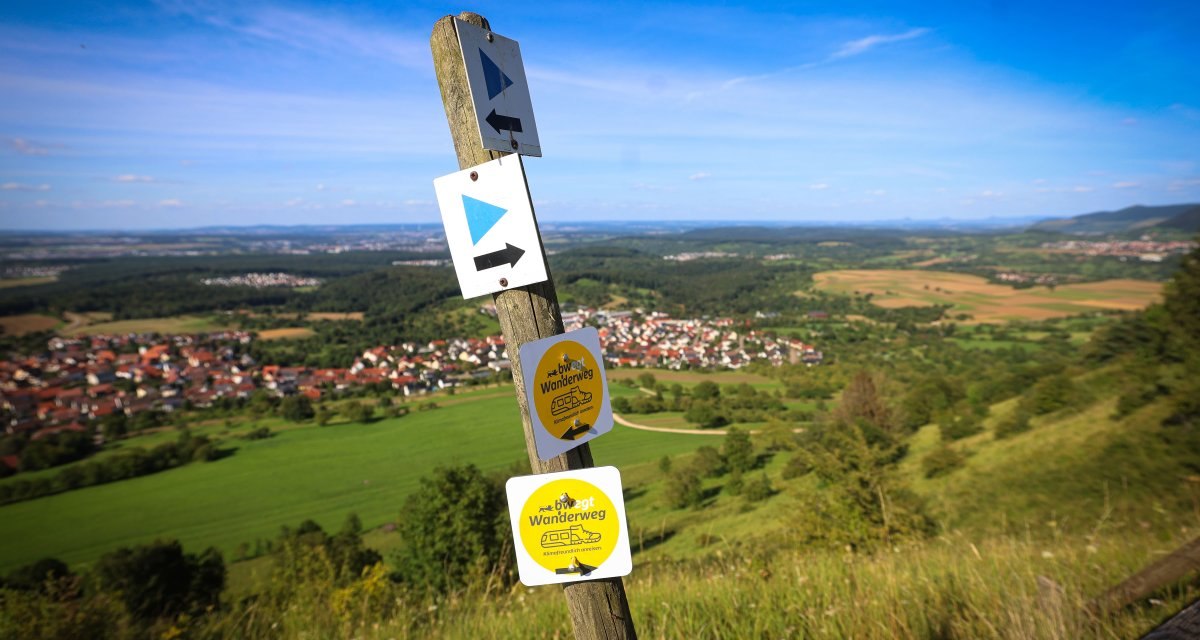 Wegweiser auf einem Hügel mit Schildern für Wanderwege, im Hintergrund eine weite Landschaft und ein Dorf unter blauem Himmel. Wegweiser auf einem Hügel mit Schildern für Wanderwege, im Hintergrund eine weite Landschaft und ein Dorf unter blauem Himmel.