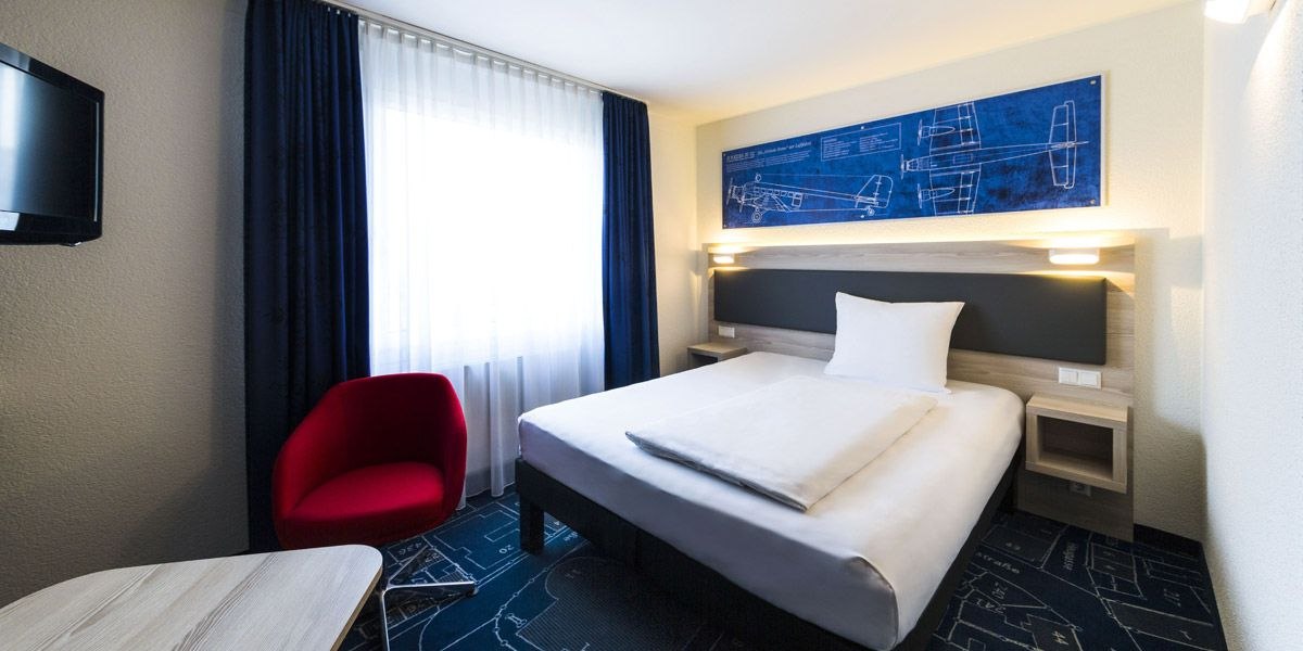 Modernes Hotelzimmer mit Einzelbett, rotem Sessel, blauem Teppich und Wandbild eines Flugzeugs. Helle Vorhänge und ein Flachbildfernseher sind vorhanden., © ibis Styles Filderstadt Messe Stuttgart Modernes Hotelzimmer mit Einzelbett, rotem Sessel, blauem Teppich und Wandbild eines Flugzeugs. Helle Vorhänge und ein Flachbildfernseher sind vorhanden., © ibis Styles Filderstadt Messe Stuttgart