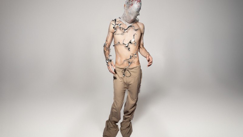 Person mit Maske und Pflanzen auf dem Oberkörper, trägt eine beige Hose in einem neutralen Raum., © Scala Kultur Live gGmbH