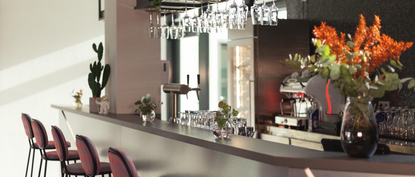 Moderne Bar mit eleganten Hockern, Pflanzen und Gläsern. Im Hintergrund eine Kaffeemaschine und Dekorationen., © Novum Management GmbH Moderne Bar mit eleganten Hockern, Pflanzen und Gläsern. Im Hintergrund eine Kaffeemaschine und Dekorationen., © Novum Management GmbH