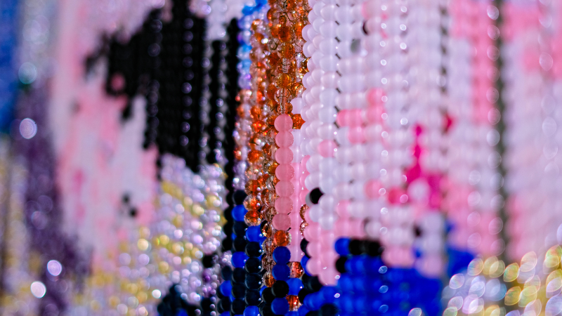 Nahaufnahme von bunten Perlen, die in verschiedenen Farben und Mustern angeordnet sind. Die Perlen erzeugen ein schimmerndes, abstraktes Muster., © Mizi Lee Nahaufnahme von bunten Perlen, die in verschiedenen Farben und Mustern angeordnet sind. Die Perlen erzeugen ein schimmerndes, abstraktes Muster., © Mizi Lee