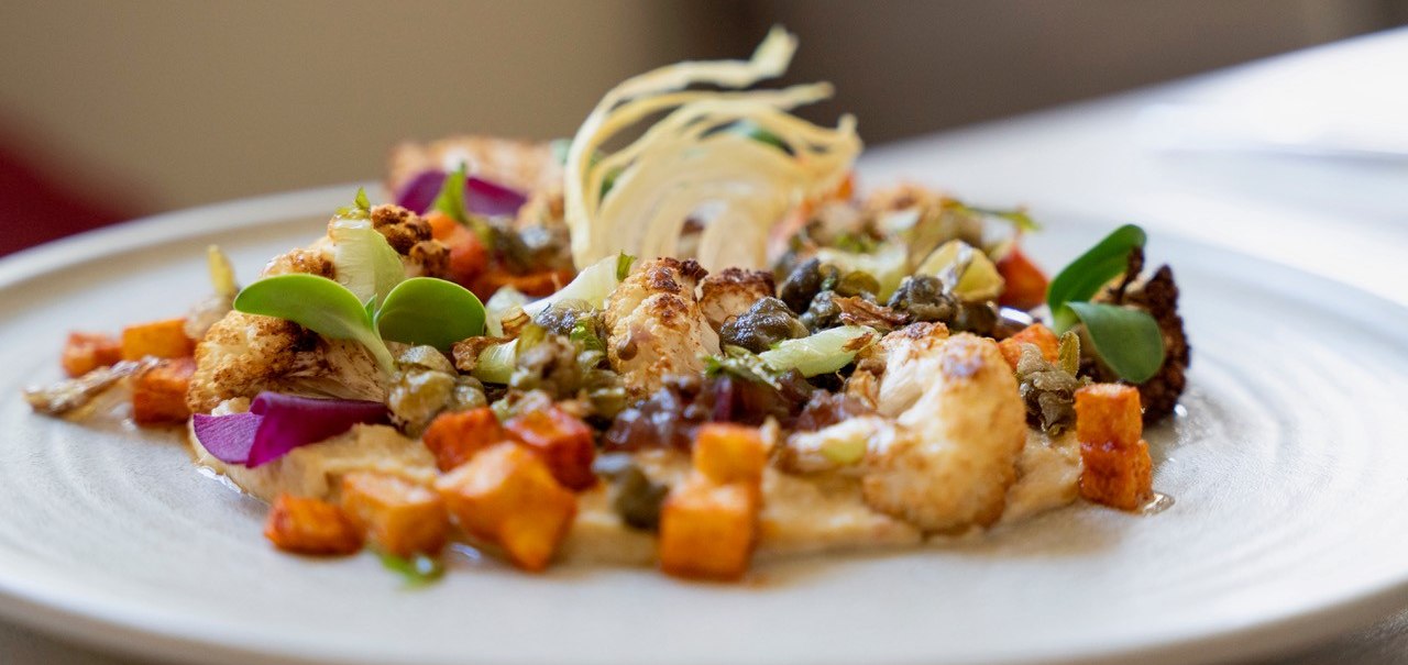 Ein Teller mit kunstvoll angerichtetem Gemüse, darunter Blumenkohl, Süßkartoffeln und Kapern, dekoriert mit grünen Blättern und einer hellen Sauce., © Veganes Gold® Ein Teller mit kunstvoll angerichtetem Gemüse, darunter Blumenkohl, Süßkartoffeln und Kapern, dekoriert mit grünen Blättern und einer hellen Sauce., © Veganes Gold®