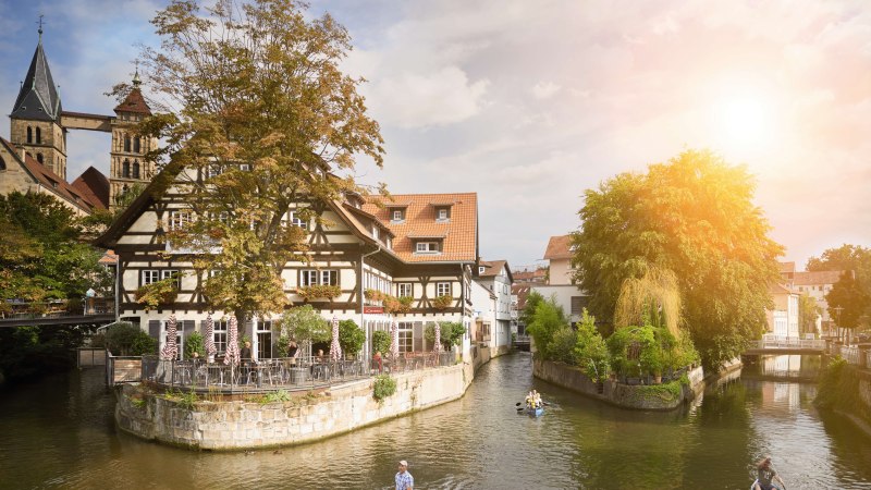 Fachwerkhaus am Roßneckarkanal, umgeben von Bäumen. Menschen sitzen im Café, während Kanufahrer auf dem Wasser unterwegs sind. Sonnenschein im Hintergrund., © Stuttgart-Marketing GmbH