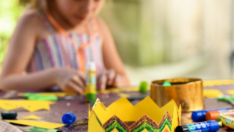Ein Kind bastelt konzentriert an einem Tisch. Im Vordergrund liegt eine bunte Papierkrone, umgeben von Bastelmaterialien wie Klebestiften und Papier.