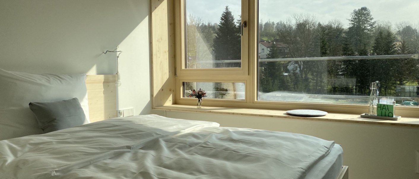 Helles Zimmer mit großem Fenster, Bett mit weißer Bettwäsche, Blick auf Bäume. Minimalistische Einrichtung mit Holzdetails., © Nina Kiesel Helles Zimmer mit großem Fenster, Bett mit weißer Bettwäsche, Blick auf Bäume. Minimalistische Einrichtung mit Holzdetails., © Nina Kiesel