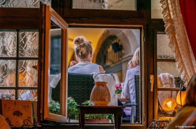 Blick durch ein offenes Fenster auf zwei Personen, die in einem gemütlichen Restaurant sitzen. Warme Beleuchtung und Dekorationen sind sichtbar., © Stuttgart Marketing GmbH Blick durch ein offenes Fenster auf zwei Personen, die in einem gemütlichen Restaurant sitzen. Warme Beleuchtung und Dekorationen sind sichtbar., © Stuttgart Marketing GmbH
