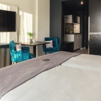 Modernes Zimmer mit großem Bett, blauen Sesseln, Astronauten-Wandbild, Flachbildfernseher und kleiner Küche., © Novum Management GmbH