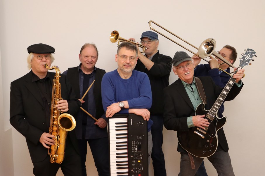 Sechs &auml;ltere M&auml;nner mit Musikinstrumenten, darunter Saxophon, Tuba, Gitarre und Keyboard, stehen vor wei&szlig;em Hintergrund., &copy; KlangWerk
