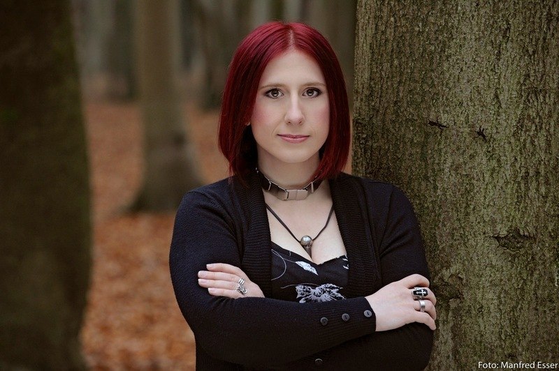 Eine Person mit roten Haaren steht mit verschr&auml;nkten Armen vor einem Baum in einem herbstlichen Wald., &copy; Scala Kultur Live gGmbH