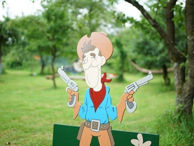Ein Cartoon-Cowboy mit zwei Pistolen steht in einem grünen Garten. Im Hintergrund sind Bäume und eine Hängematte zu sehen., © Cool-Tours StattReisen Ein Cartoon-Cowboy mit zwei Pistolen steht in einem grünen Garten. Im Hintergrund sind Bäume und eine Hängematte zu sehen., © Cool-Tours StattReisen