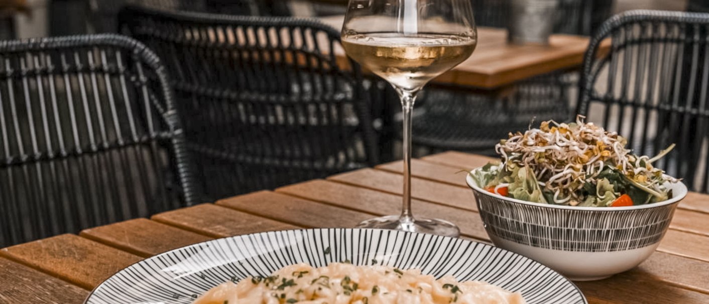 Ein Glas Weißwein, ein Teller Pasta und ein Salat auf einem Holztisch in einem Restaurant mit geflochtenen Stühlen., © SMG, itchyfeet-travel.de Ein Glas Weißwein, ein Teller Pasta und ein Salat auf einem Holztisch in einem Restaurant mit geflochtenen Stühlen., © SMG, itchyfeet-travel.de