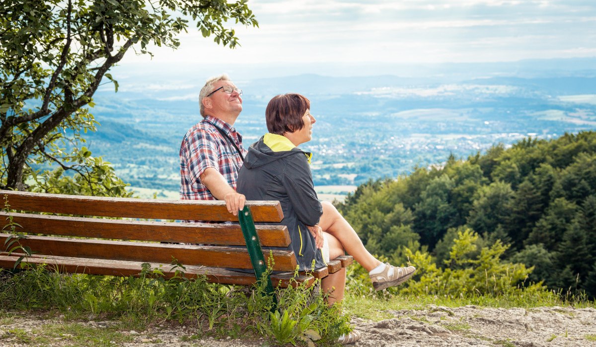 Ein älteres Paar sitzt auf einer Bank und genießt die Aussicht auf eine weite, grüne Landschaft unter einem bewölkten Himmel., © hochgehberge