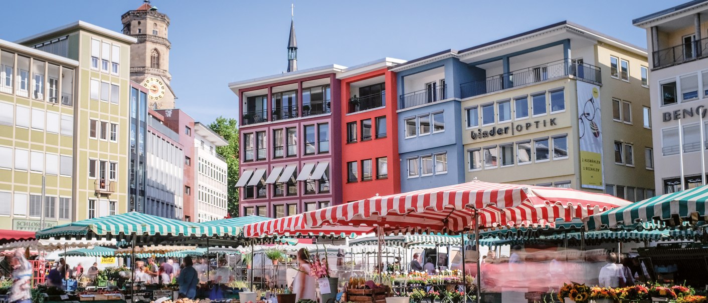 Wochenmarkt in Stuttgart mit bunten Marktständen und Gebäuden im Hintergrund. Menschen schlendern zwischen den Ständen, während die Sonne scheint., © SMG, trickytine