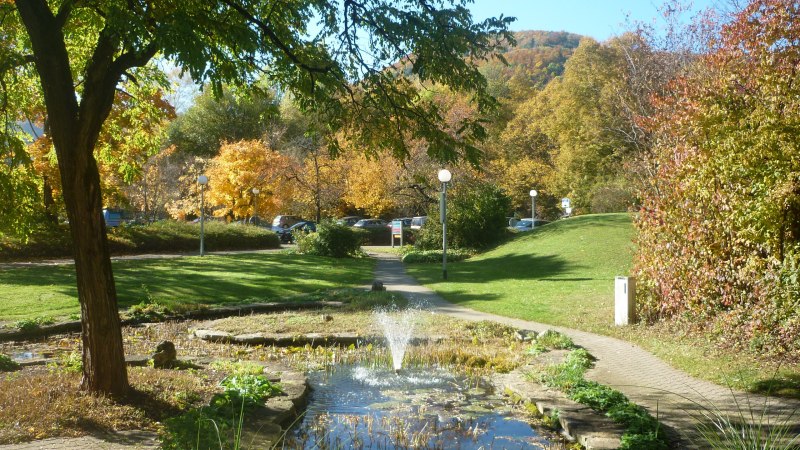 Ein herbstlicher Park mit bunten B&auml;umen, einem kleinen Teich mit Springbrunnen und einem gepflasterten Weg. Die Sonne scheint hell am blauen Himmel.
