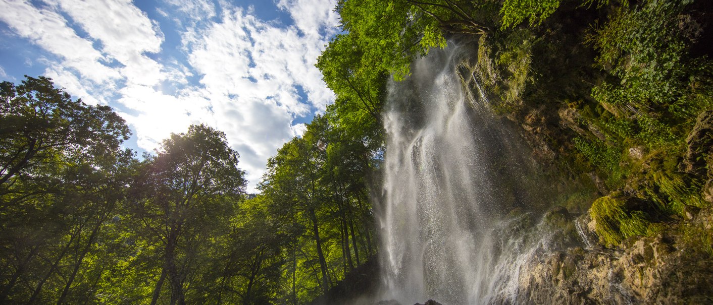 Der Uracher Wasserfall stürzt über Felsen, umgeben von grünen Bäumen und einem blauen Himmel mit Wolken., © Bad Urach Tourismus Der Uracher Wasserfall stürzt über Felsen, umgeben von grünen Bäumen und einem blauen Himmel mit Wolken., © Bad Urach Tourismus