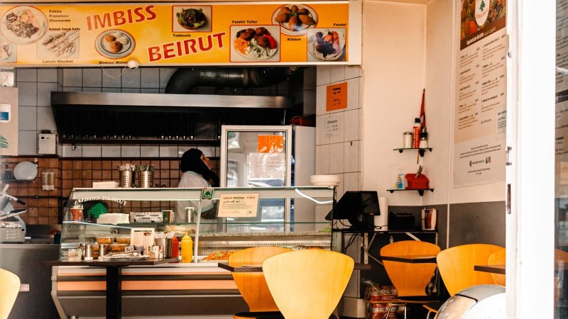Ein Imbiss mit gelbem Schild 'Imbiss Beirut', leeren Tischen und einer Person hinter der Theke., © SMG, Sarah Schmid