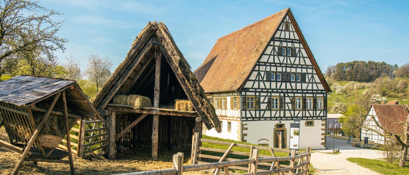 Fachwerkhaus und Scheune im Freilichtmuseum Beuren, umgeben von blühenden Bäumen und grünen Hügeln unter blauem Himmel., © Stuttgart-Marketing GmbH, Sarah Schmid